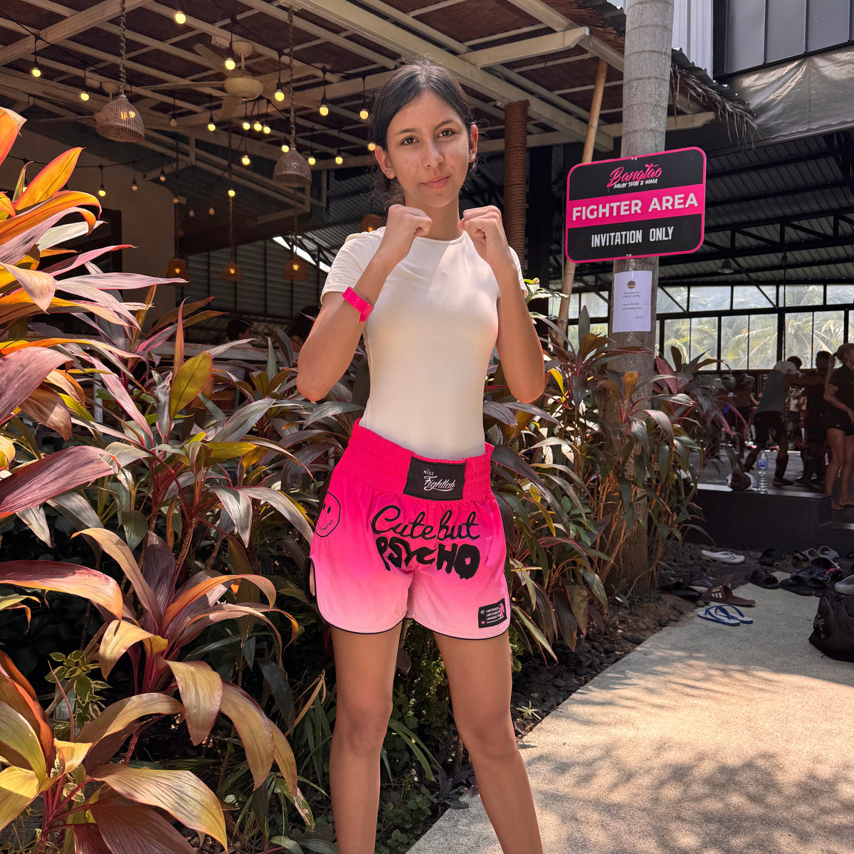 A woman posing in a tropical outdoor setting, wearing bright pink and white gradient athletic shorts with 'Celebit Arena' printed on the front, paired with a white sleeveless top.