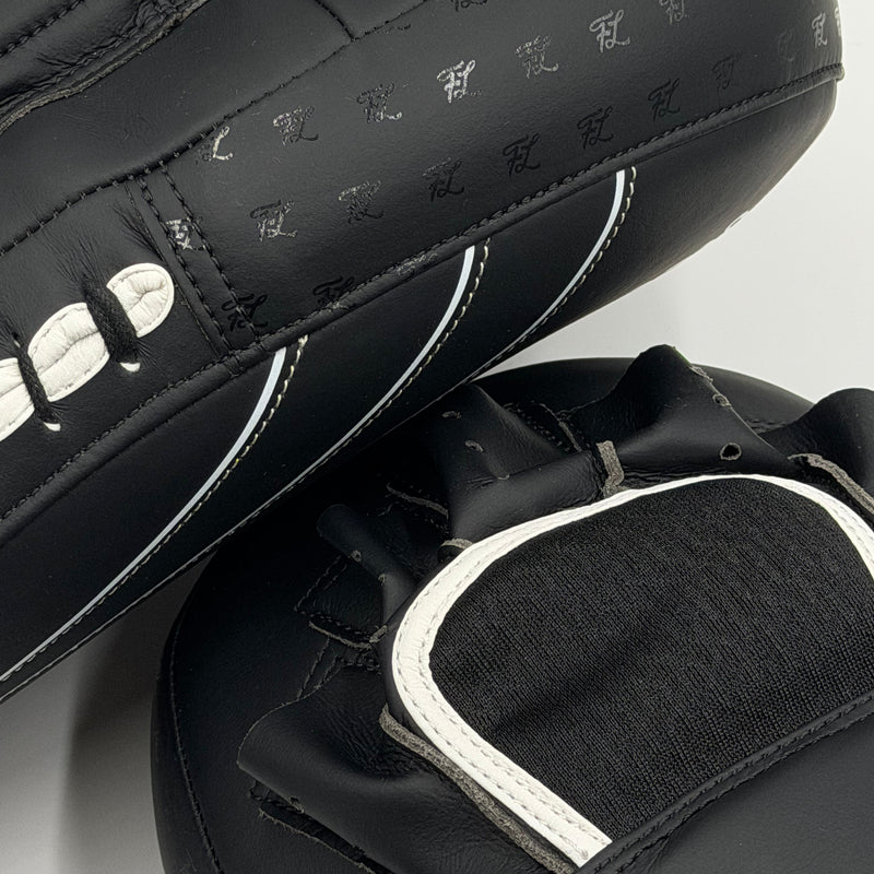 Black leather martial arts kicking pads with white contrast stitching and white logo lettering, shown in a close-up studio shot.