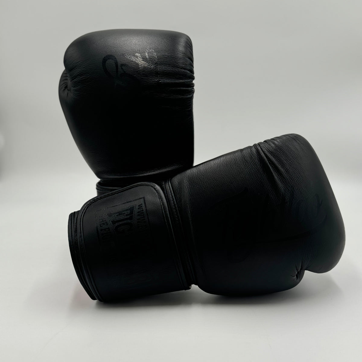 A pair of black leather boxing gloves, one standing upright and the other lying on its side, displayed on a light gray studio background.