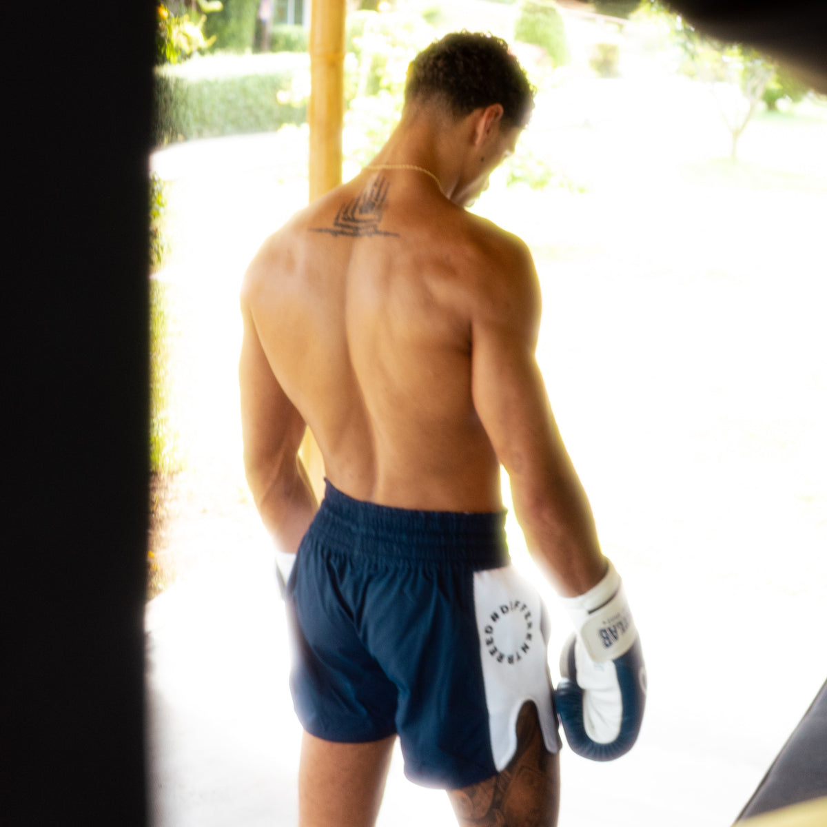 A shirtless man viewed from behind wearing navy blue athletic shorts and white boxing gloves, standing in a doorway with a bright outdoor background.