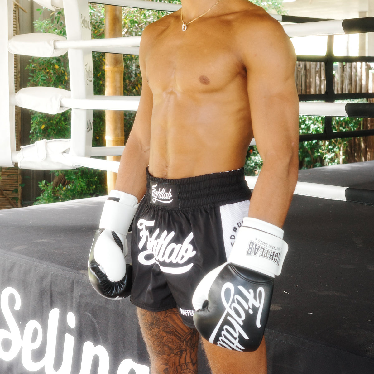 A shirtless man standing in a boxing ring wearing black and white Fightlab boxing shorts and gloves, with a thatched-roof structure in the background.