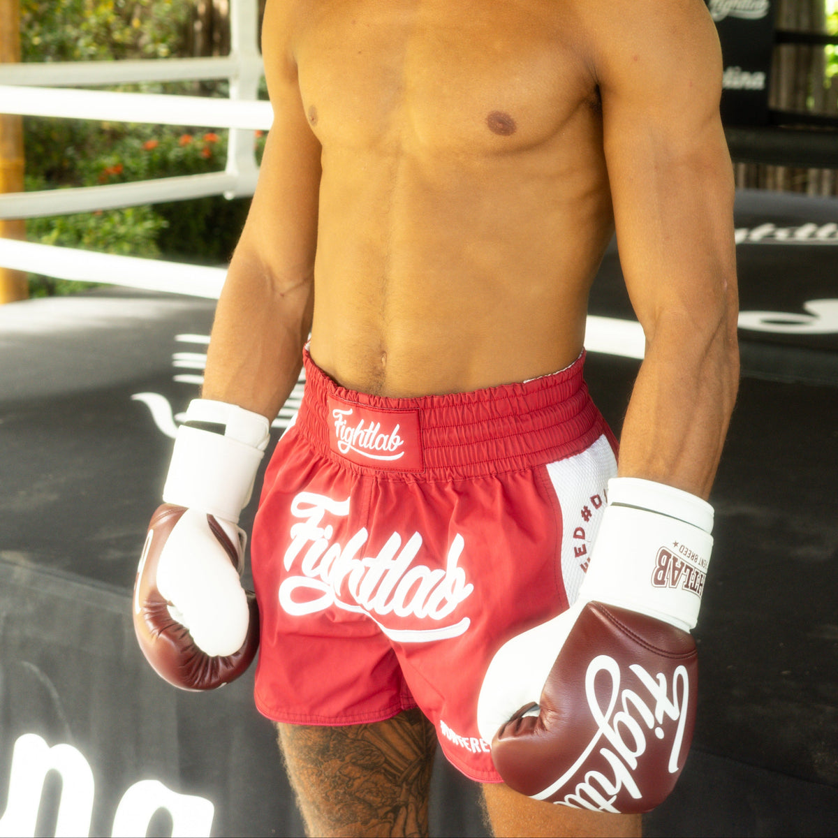A shirtless man wearing red and white Muay Thai shorts and matching gloves stands in a boxing ring under a wooden roof structure. The brand name 'Flykladd' is clearly visible on the shorts and gloves.