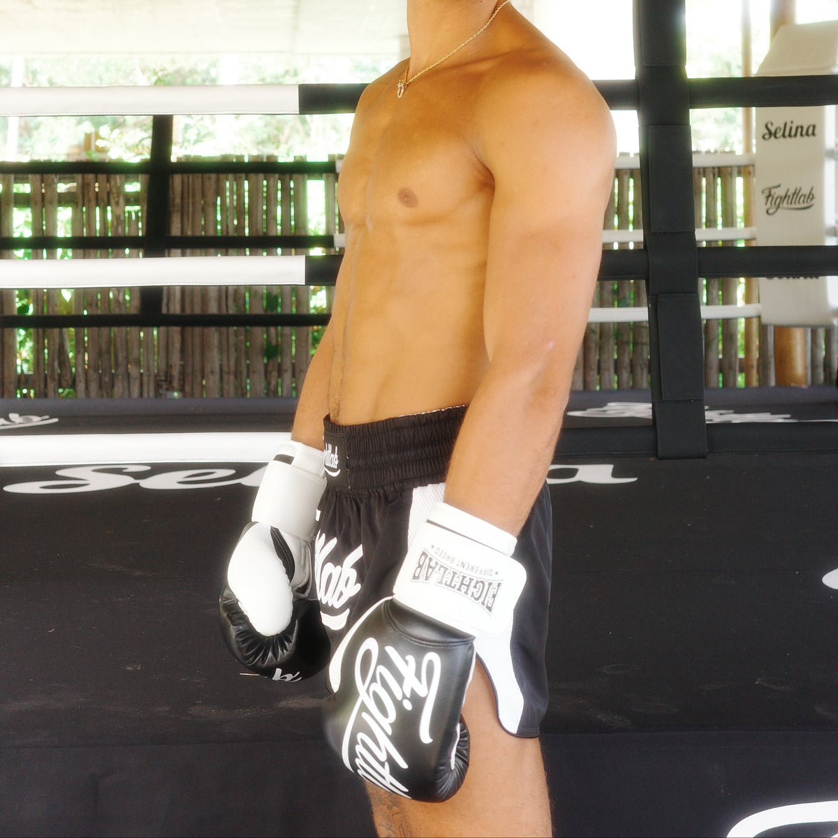 A shirtless man wearing black and white boxing fightlab  gloves and black athletic shorts stands inside a boxing ring under a thatched roof structure. The gloves show the brand name 'Standard'.