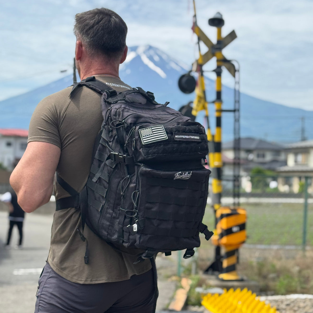 A man viewed from behind wearing a large black tactical backpack with multiple compartments and an American flag patch, standing outdoors in front of a mountain and railroad crossing.
